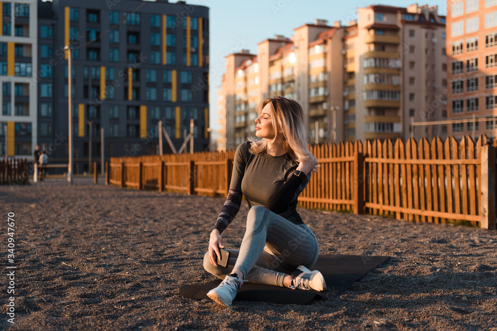 Sporty young woman sitting on yoga mat near playground with closed eyes, holding cellphone in her hand. The girl enjoys the sunset after an active workout