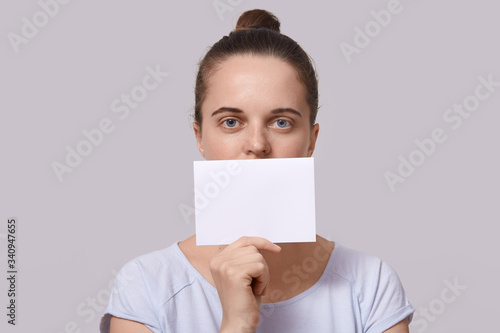 Horizontal photo of good looking serious cute young lady looking directly at camera, holding blank sheet of paper for message, having peaceful facial expression. Copyspace for advertisement.