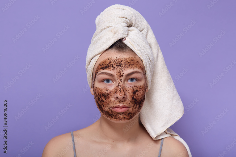 Horizontal shot of serious attractive young female looking directly at camera, having applied coffee scrub on face, cleaning skin, having towel on head, home beauty procedures. Beauty concept. Horizontal shot of serious attractive young female looking directly at camera, having applied coffee scrub on face, cleaning skin, having towel on head, home beauty procedures. Beauty concept.