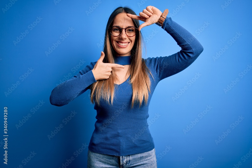 Young beautiful blonde woman with blue eyes wearing glasses standing over blue background smiling making frame with hands and fingers with happy face. Creativity and photography concept. Young beautiful blonde woman with blue eyes wearing glasses standing over blue background smiling making frame with hands and fingers with happy face. Creativity and photography concept.