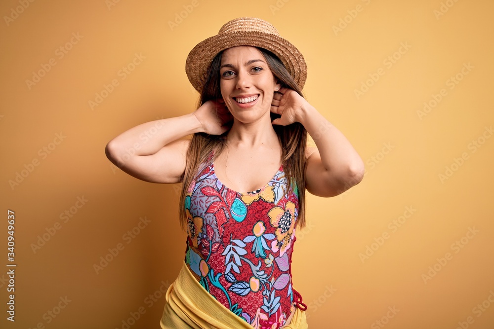 Young beautiful blonde woman wearing swimsuit and summer hat over yellow background relaxing and stretching, arms and hands behind head and neck smiling happy Young beautiful blonde woman wearing swimsuit and summer hat over yellow background relaxing and stretching, arms and hands behind head and neck smiling happy