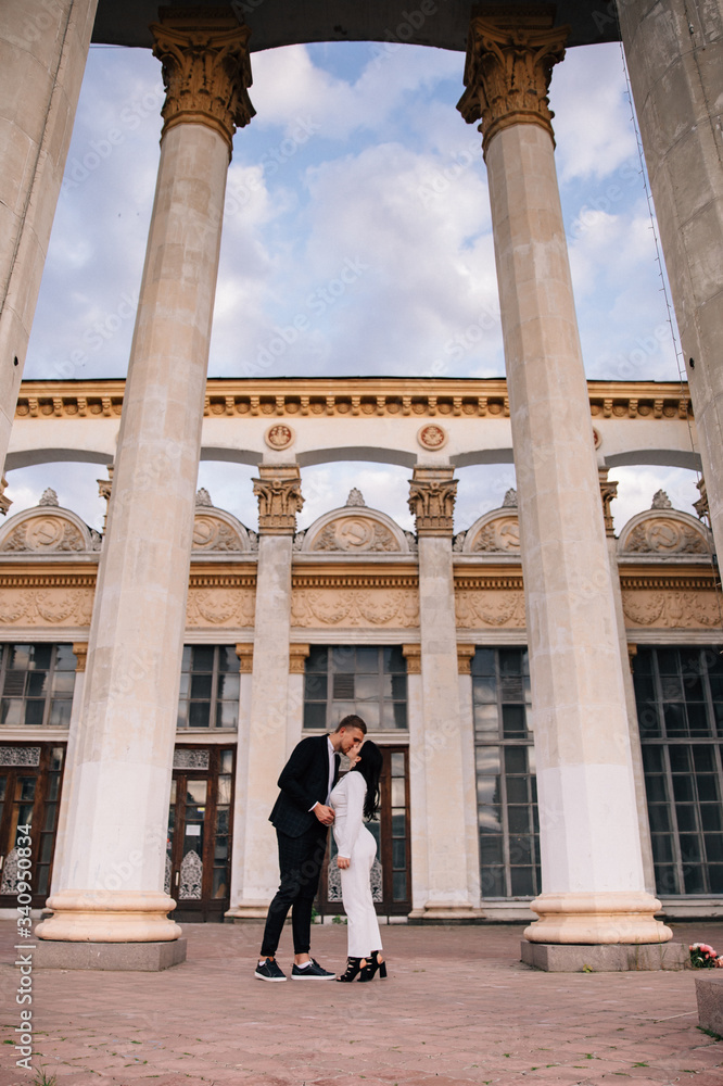 bride and groom in a white pantsuit kiss between large white columns bride and groom in a white pantsuit kiss between large white columns
