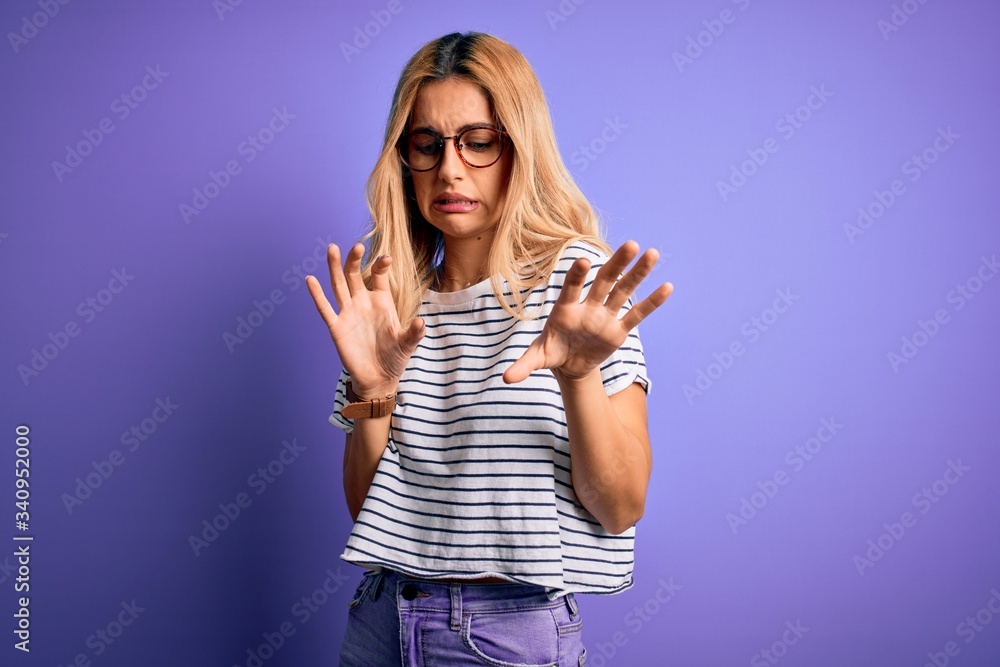 Fototapeta premium Young beautiful blonde woman wearing striped t-shirt and glasses over purple background disgusted expression, displeased and fearful doing disgust face because aversion reaction.