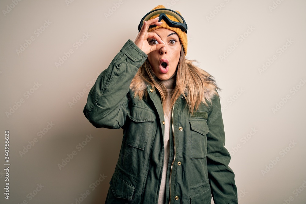 Young brunette skier woman wearing snow clothes and ski goggles over white background doing ok gesture shocked with surprised face, eye looking through fingers. Unbelieving expression. Young brunette skier woman wearing snow clothes and ski goggles over white background doing ok gesture shocked with surprised face, eye looking through fingers. Unbelieving expression.