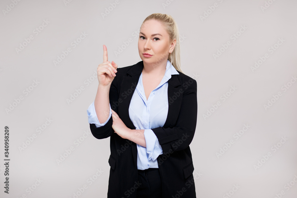 portrait of beautiful plus size blonde woman in black business suit showing idea or attention sign over gray background