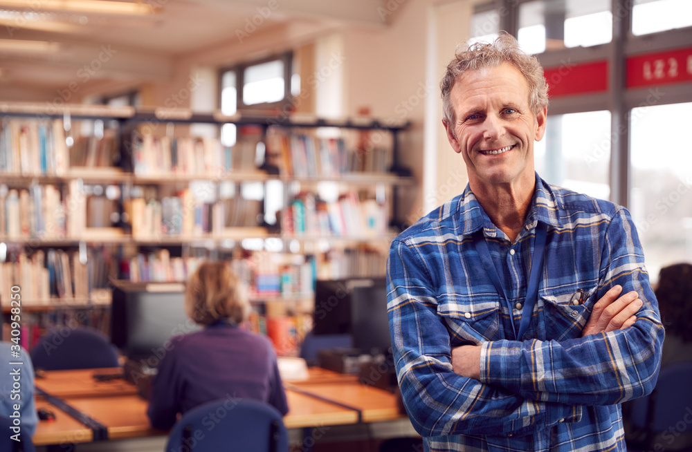 Portrait Of Mature Male Teacher Or Student In Library With Other Students Studying In Background Portrait Of Mature Male Teacher Or Student In Library With Other Students Studying In Background