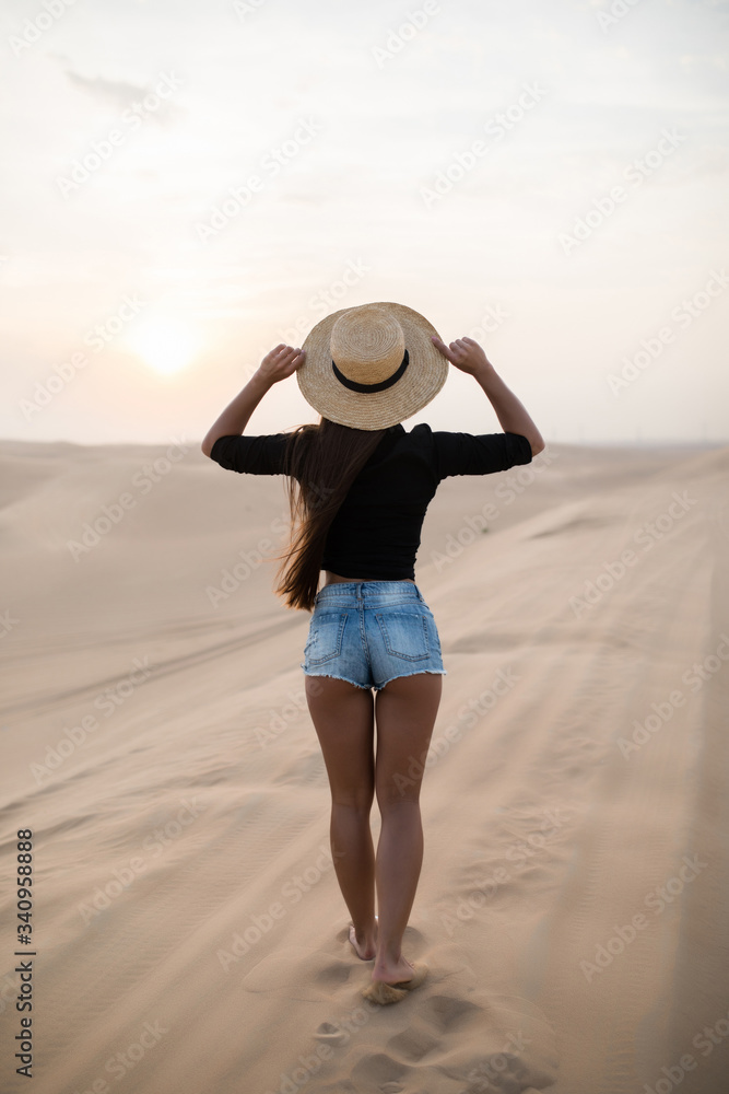 Young sexy woman with straw hat walking barefoot on desert dunes at sunset