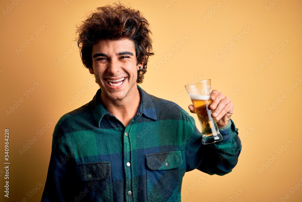 Young handsome man smiling happy. Standing with smile on face drinking glass of beer over isolated yellow background Young handsome man smiling happy. Standing with smile on face drinking glass of beer over isolated yellow background