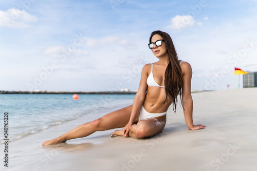 An attractive young woman wearing a white bikini sits on a beach with her elbow on her knee in the background.