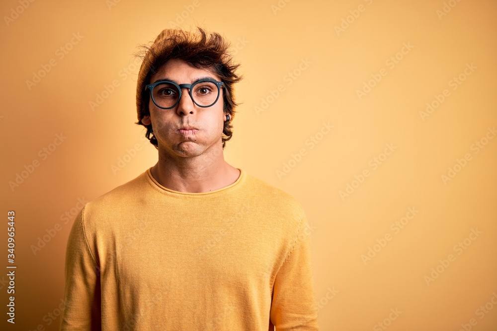 Young handsome man wearing casual t-shirt and glasses over isolated yellow background puffing cheeks with funny face. Mouth inflated with air, crazy expression. Young handsome man wearing casual t-shirt and glasses over isolated yellow background puffing cheeks with funny face. Mouth inflated with air, crazy expression.