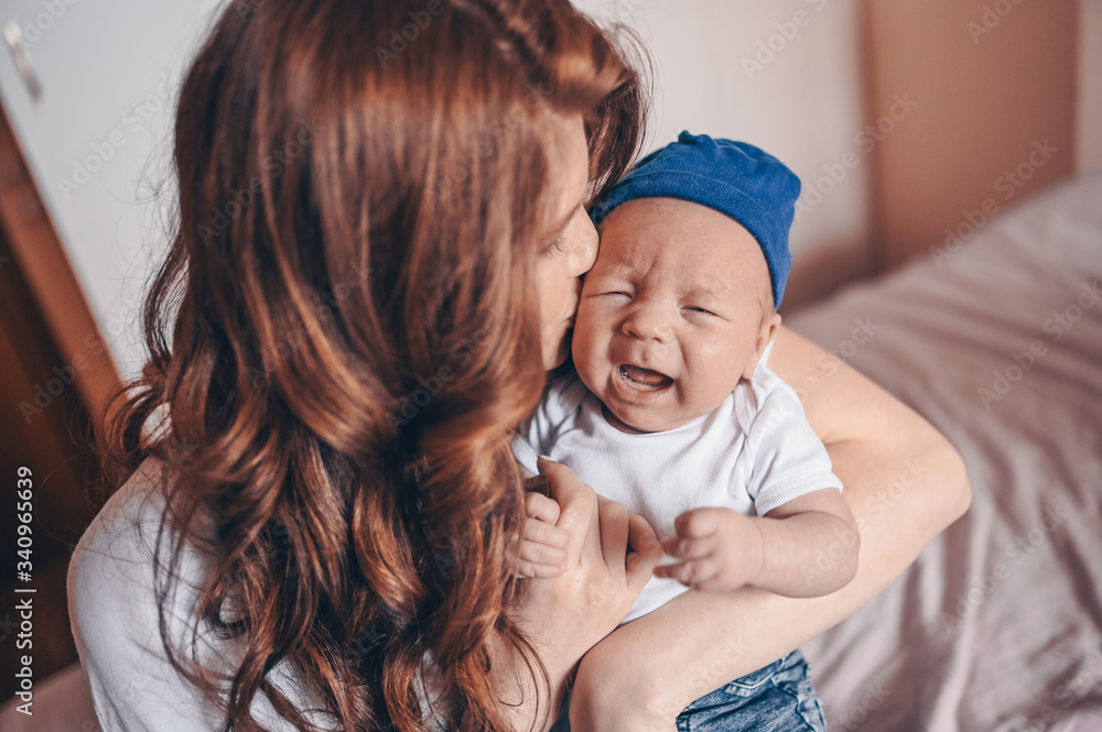 Close up portrait of happy young mom holding her crying baby in blue jeans and white t-shirt and cap. Young happy family, mother playing with cute emotional little newborn child son in the bedroom. Close up portrait of happy young mom holding her crying baby in blue jeans and white t-shirt and cap. Young happy family, mother playing with cute emotional little newborn child son in the bedroom.