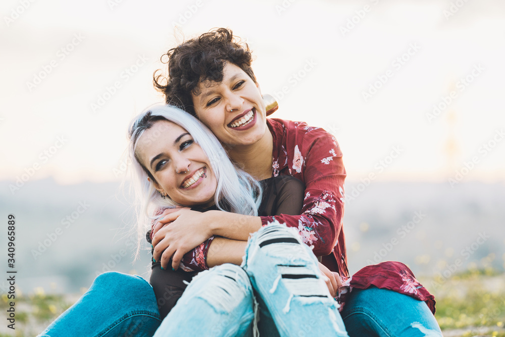 Laughing close girlfriends in stylish casual outfits sitting embracing each other on green grass with sky on background Laughing close girlfriends in stylish casual outfits sitting embracing each other on green grass with sky on background