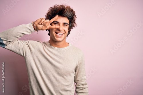 Young handsome man wearing casual t-shirt standing over isolated pink background Doing peace symbol with fingers over face, smiling cheerful showing victory