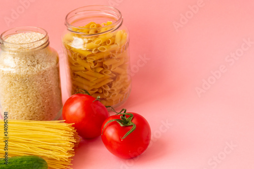 Noodles, rice, pasta in glass jars stand on a pink background. Nearby are red tomatoes and green cucumber. Raw materials for cooking.