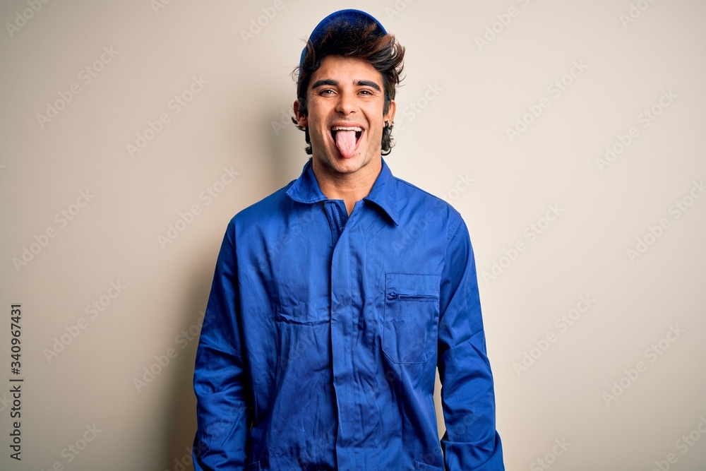 Young mechanic man wearing blue cap and uniform standing over isolated white background sticking tongue out happy with funny expression. Emotion concept. Young mechanic man wearing blue cap and uniform standing over isolated white background sticking tongue out happy with funny expression. Emotion concept.