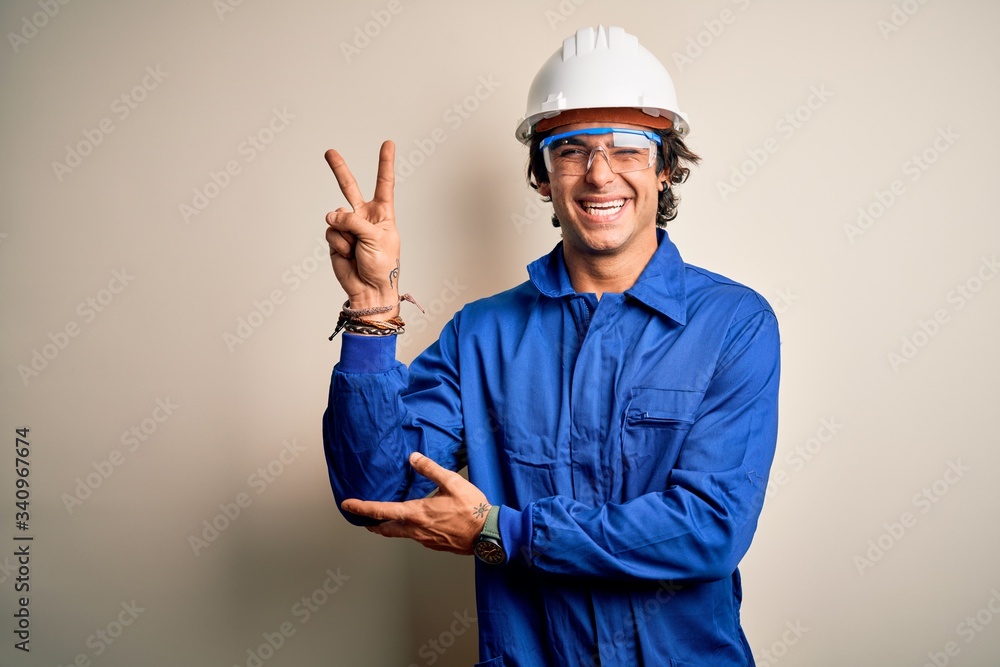 Young constructor man wearing uniform and security helmet over isolated white background smiling with happy face winking at the camera doing victory sign. Number two. Young constructor man wearing uniform and security helmet over isolated white background smiling with happy face winking at the camera doing victory sign. Number two.