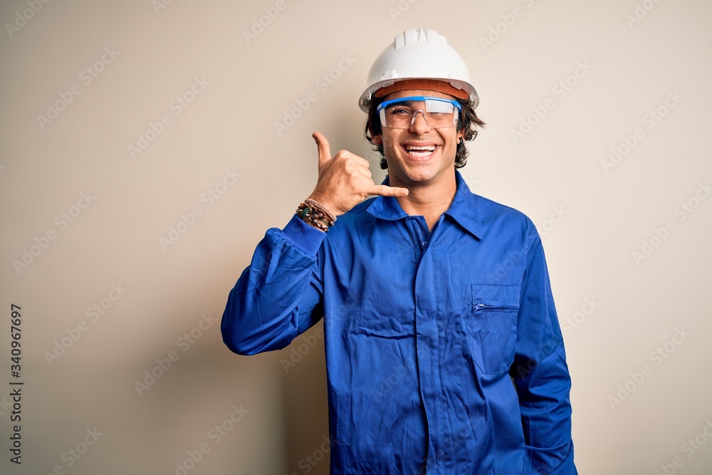 Young constructor man wearing uniform and security helmet over isolated white background smiling doing phone gesture with hand and fingers like talking on the telephone. Communicating concepts.