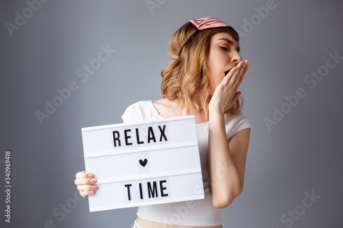Beautiful woman dressed in white t-shirt and sleep head band on her hair is yawning. Woman has a panel wih text: relax time. 