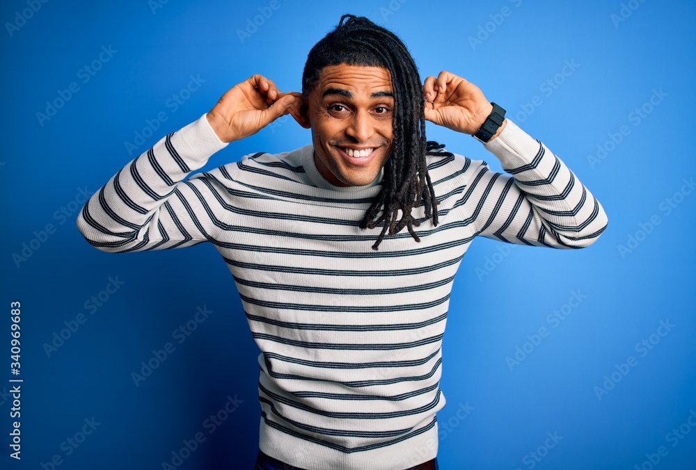 Naklejka premium Young handsome african american afro man with dreadlocks wearing casual striped sweater Smiling pulling ears with fingers, funny gesture. Audition problem