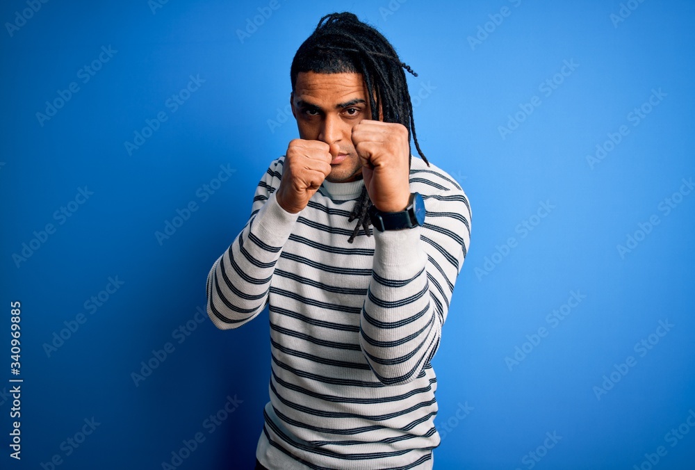 Young handsome african american afro man with dreadlocks wearing casual striped sweater Ready to fight with fist defense gesture, angry and upset face, afraid of problem Young handsome african american afro man with dreadlocks wearing casual striped sweater Ready to fight with fist defense gesture, angry and upset face, afraid of problem