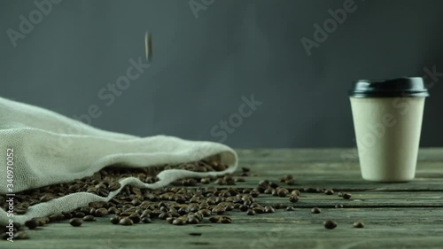 coffee beans fallings on a wooden table beside a beige bag full of coffee beans along side a coffe cup in studio