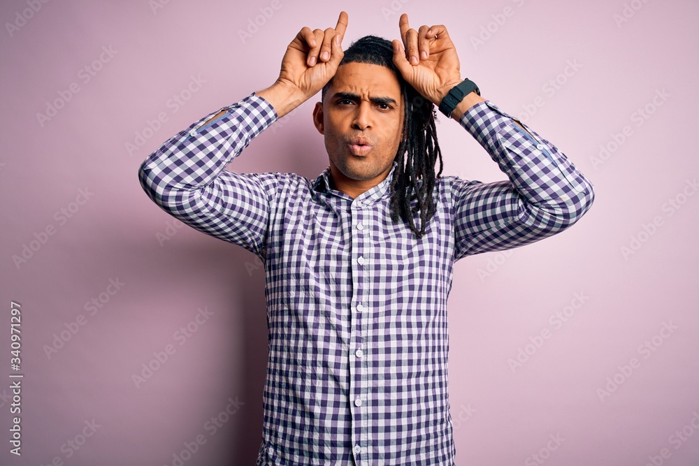 Young handsome african american afro man with dreadlocks wearing casual shirt doing funny gesture with finger over head as bull horns