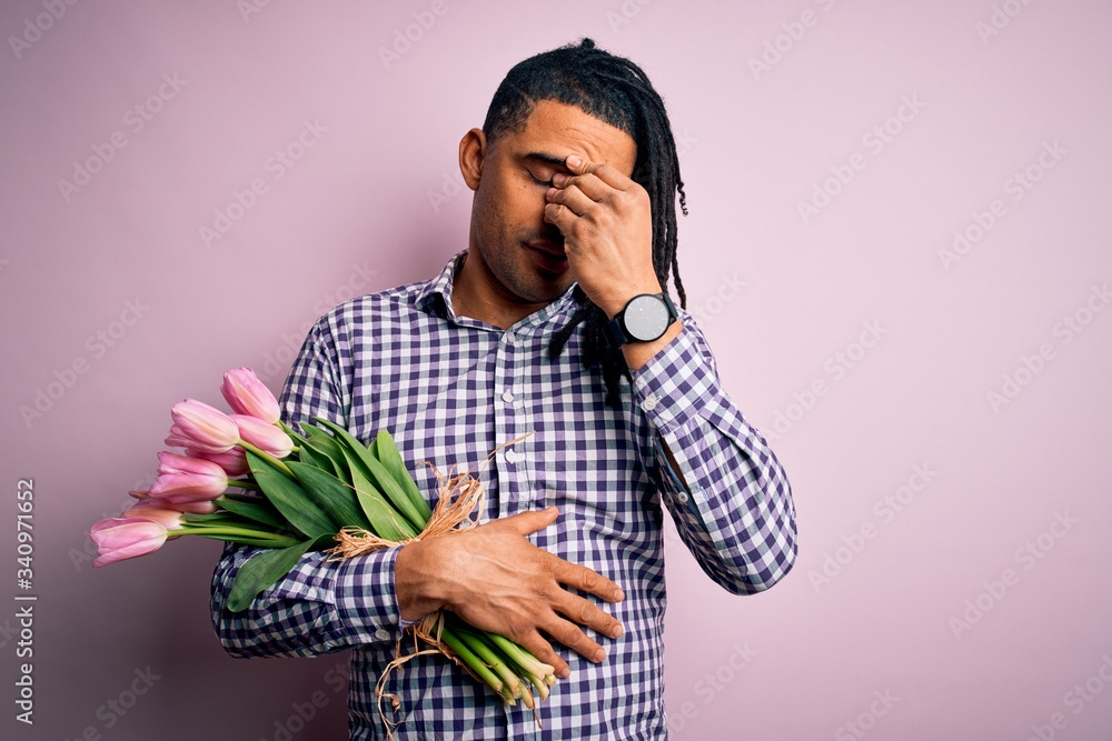 Young african american afro romantic man with dreadlocks holding bouquet of pink tulips tired rubbing nose and eyes feeling fatigue and headache. Stress and frustration concept. Young african american afro romantic man with dreadlocks holding bouquet of pink tulips tired rubbing nose and eyes feeling fatigue and headache. Stress and frustration concept.