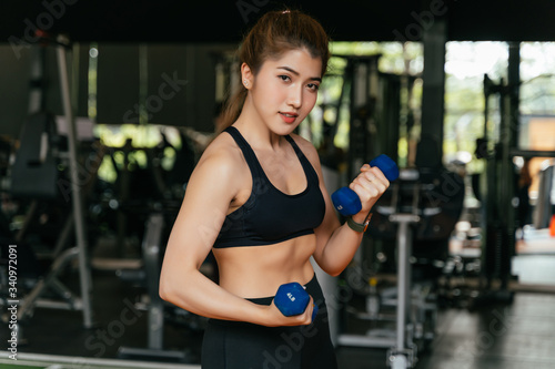 Smiling Asian athletic woman looks at camera while working out with dumbells at fitness gym. One happy young female athlete practicing weightlifting.