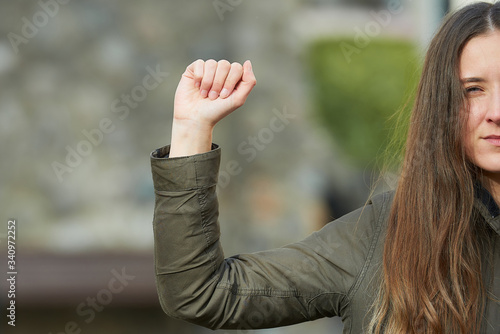 A half-part portrait of a young woman with long hair in a green jacket. A girl in an old street shows a fist as a symbol of the fight. 