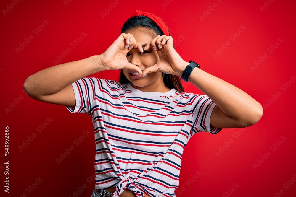 Young beautiful asian girl wearing casual striped t-shirt over isolated red background Doing heart shape with hand and fingers smiling looking through sign Young beautiful asian girl wearing casual striped t-shirt over isolated red background Doing heart shape with hand and fingers smiling looking through sign