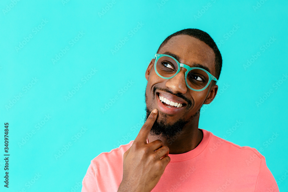 © Carlos David - Portrait of a happy young man with glasses smiling, thinking and looking up with finger on chin, against blue studio background © Carlos David - Portrait of a happy young man with glasses smiling, thinking and looking up with finger on chin, against blue studio background