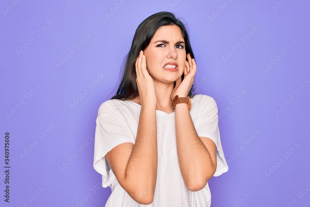 Young beautiful brunette woman wearing casual white t-shirt over purple background covering ears with fingers with annoyed expression for the noise of loud music. Deaf concept. Young beautiful brunette woman wearing casual white t-shirt over purple background covering ears with fingers with annoyed expression for the noise of loud music. Deaf concept.