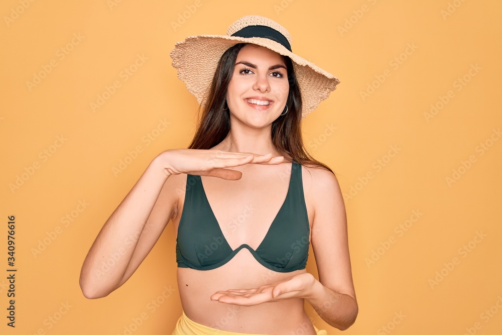 Young beautiful girl wearing swimwear bikini and summer sun hat over yellow background gesturing with hands showing big and large size sign, measure symbol. Smiling looking at the camera. Measuring Young beautiful girl wearing swimwear bikini and summer sun hat over yellow background gesturing with hands showing big and large size sign, measure symbol. Smiling looking at the camera. Measuring