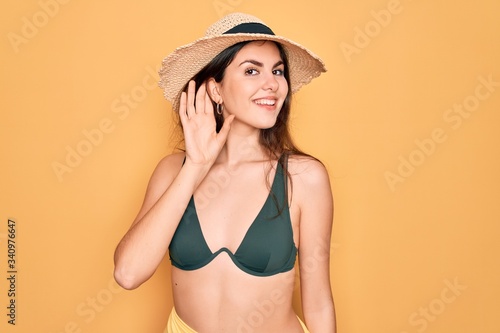 Young beautiful girl wearing swimwear bikini and summer sun hat over yellow background smiling with hand over ear listening an hearing to rumor or gossip. Deafness concept.