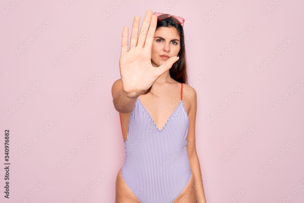 Young beautiful fashion girl wearing swimwear swimsuit and sunglasses over pink background doing stop sing with palm of the hand. Warning expression with negative and serious gesture on the face. Young beautiful fashion girl wearing swimwear swimsuit and sunglasses over pink background doing stop sing with palm of the hand. Warning expression with negative and serious gesture on the face.