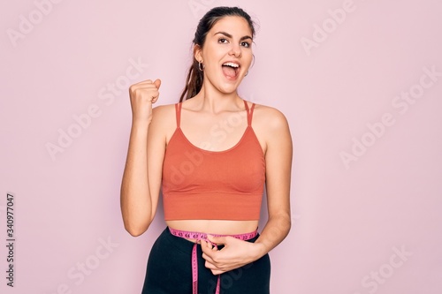 Young beautiful fitness woman wearing sport excersie clothes using measuring tape screaming proud and celebrating victory and success very excited, cheering emotion © Krakenimages.com