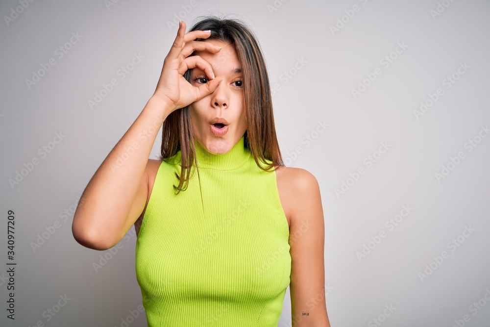 Young beautiful brunette girl wearing casual summer t-shirt over isolated white background doing ok gesture shocked with surprised face, eye looking through fingers. Unbelieving expression. Young beautiful brunette girl wearing casual summer t-shirt over isolated white background doing ok gesture shocked with surprised face, eye looking through fingers. Unbelieving expression.
