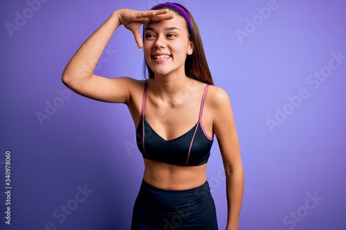 Young beautiful sporty girl doing sport wearing sportswear over isolated purple background very happy and smiling looking far away with hand over head. Searching concept. © Krakenimages.com