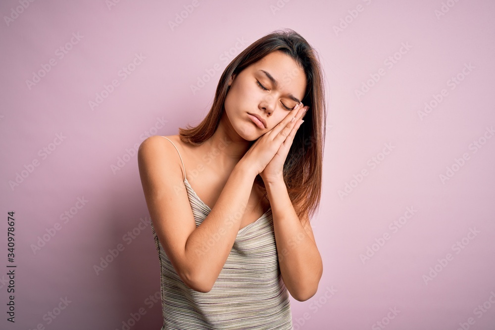 Young beautiful brunette girl wearing casual striped t-shirt over isolated pink background sleeping tired dreaming and posing with hands together while smiling with closed eyes. Young beautiful brunette girl wearing casual striped t-shirt over isolated pink background sleeping tired dreaming and posing with hands together while smiling with closed eyes.