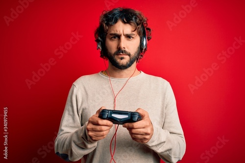 Young gamer man with curly hair and beard playing video game using joystick and headphones with a confident expression on smart face thinking serious