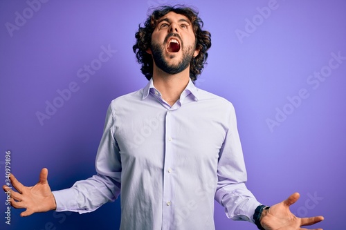 Young handsome business man with beard wearing shirt standing over purple background crazy and mad shouting and yelling with aggressive expression and arms raised. Frustration concept.