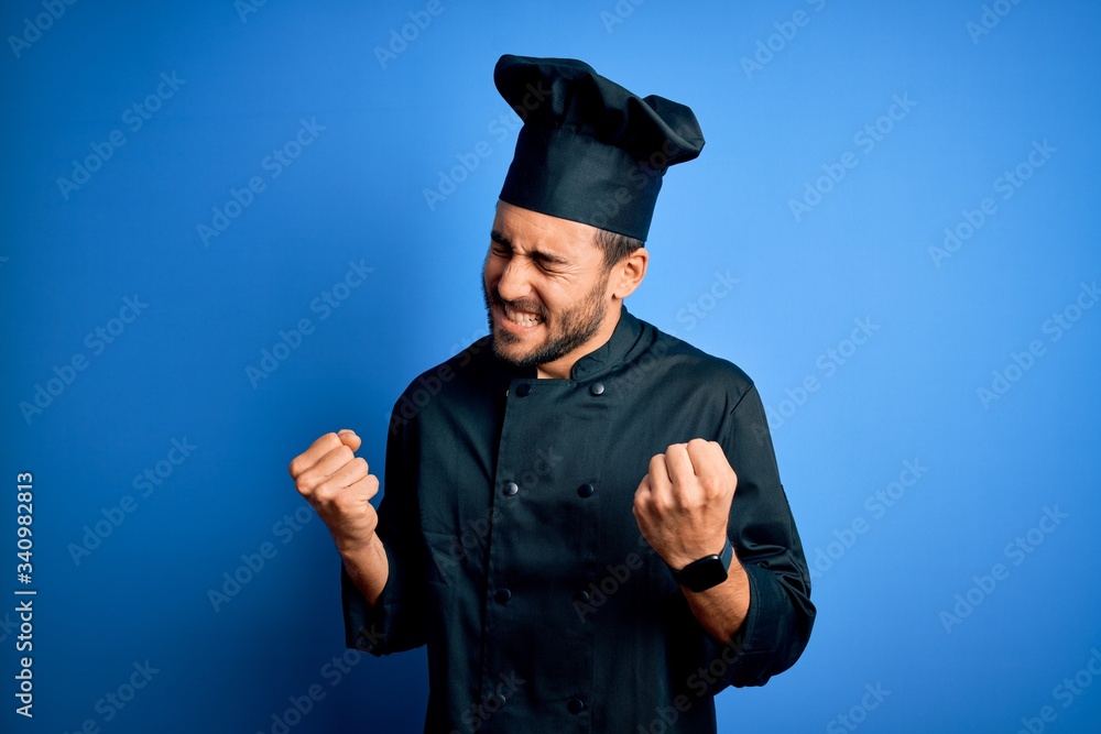 Young handsome chef man with beard wearing cooker uniform and hat over blue background very happy and excited doing winner gesture with arms raised, smiling and screaming for success. Celebration Young handsome chef man with beard wearing cooker uniform and hat over blue background very happy and excited doing winner gesture with arms raised, smiling and screaming for success. Celebration
