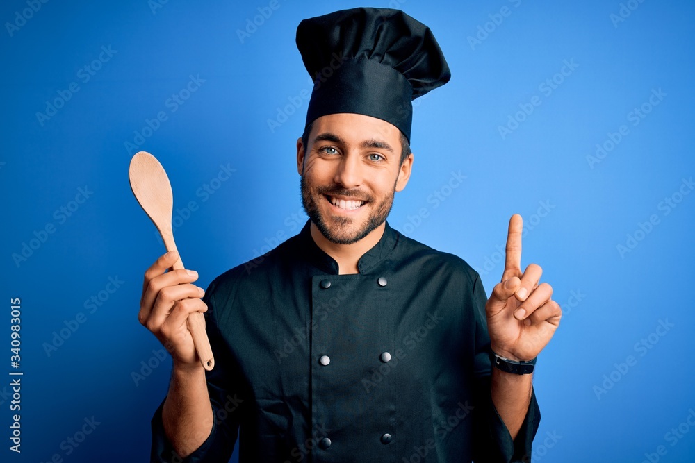 Fototapeta premium Young cooker man with beard wearing uniform holding wooden spoon over blue background surprised with an idea or question pointing finger with happy face, number one