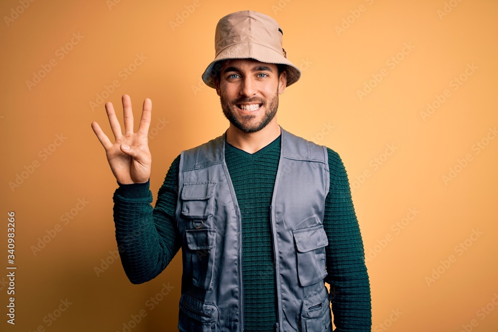 Handsome tourist man with beard on vacation wearing explorer hat over yellow background showing and pointing up with fingers number four while smiling confident and happy. Handsome tourist man with beard on vacation wearing explorer hat over yellow background showing and pointing up with fingers number four while smiling confident and happy.
