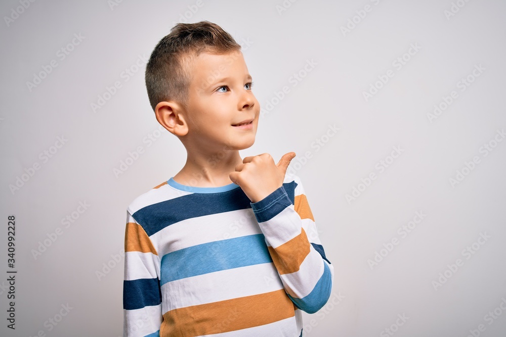 Young little caucasian kid with blue eyes standing wearing striped shirt over isolated background smiling with happy face looking and pointing to the side with thumb up. Young little caucasian kid with blue eyes standing wearing striped shirt over isolated background smiling with happy face looking and pointing to the side with thumb up.