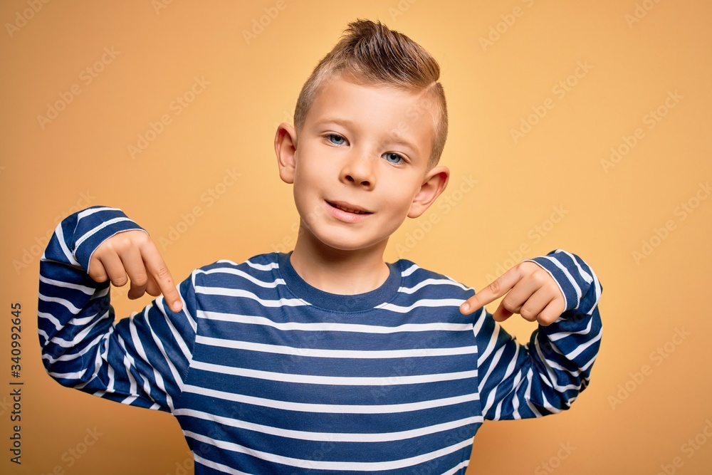 Young little caucasian kid with blue eyes wearing nautical striped shirt over yellow background looking confident with smile on face, pointing oneself with fingers proud and happy.