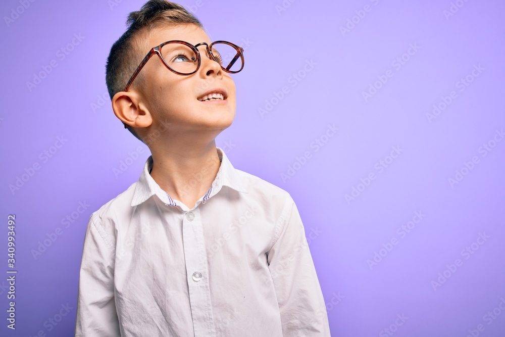 Young little caucasian kid with blue eyes wearing glasses and white shirt over purple background looking away to side with smile on face, natural expression. Laughing confident.