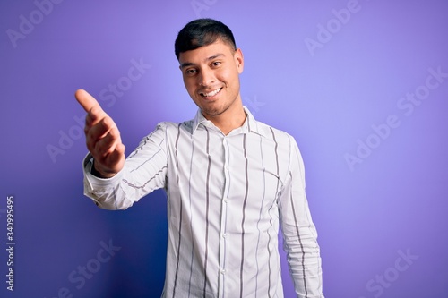 Young handsome hispanic man wearing elegant business shirt standing over purple background smiling friendly offering handshake as greeting and welcoming. Successful business.