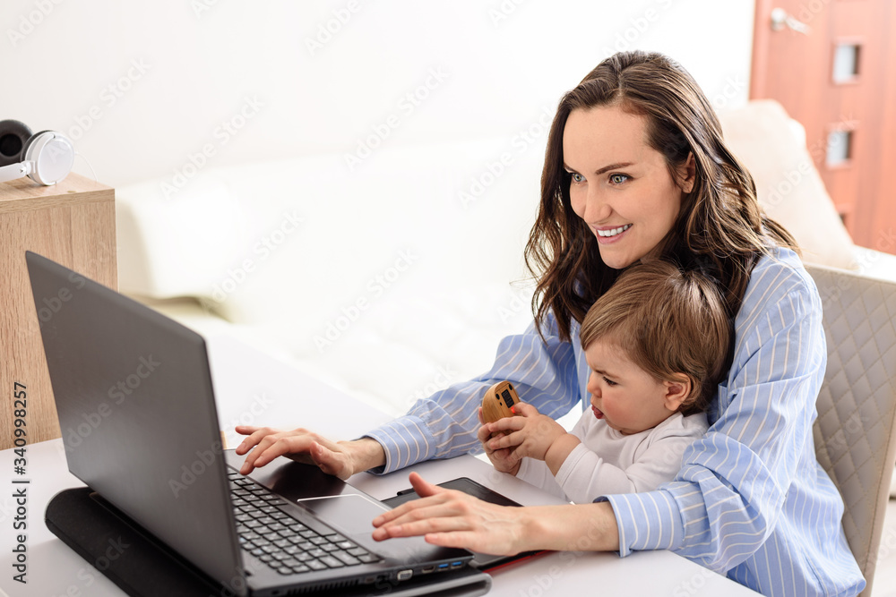 young smiling mother in blue shirt working on laptop with her baby at home, working on maternity leave young smiling mother in blue shirt working on laptop with her baby at home, working on maternity leave