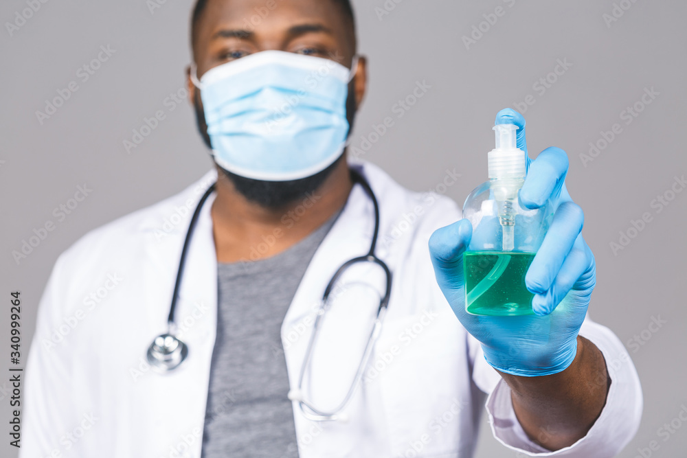 African american doctor man in gown face mask gloves isolated on grey background. Epidemic pandemic coronavirus 2019-ncov sars covid-19 flu virus. Holding bottle liquid antibacterial sanitizer. African american doctor man in gown face mask gloves isolated on grey background. Epidemic pandemic coronavirus 2019-ncov sars covid-19 flu virus. Holding bottle liquid antibacterial sanitizer.